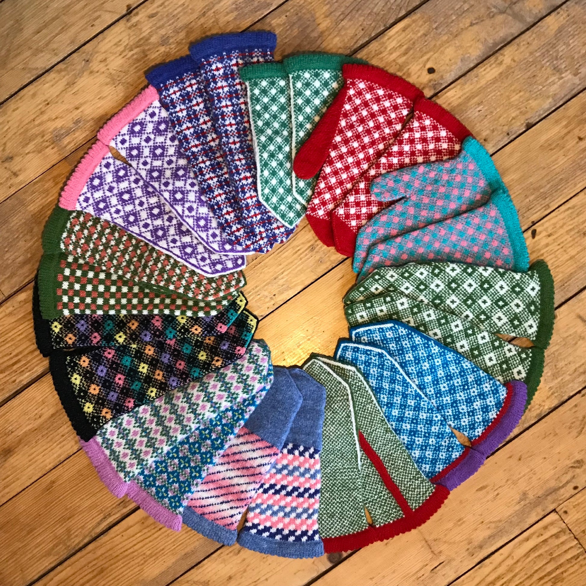 18 different colorful Fair Isle mittens in a circle, on a wood floor
